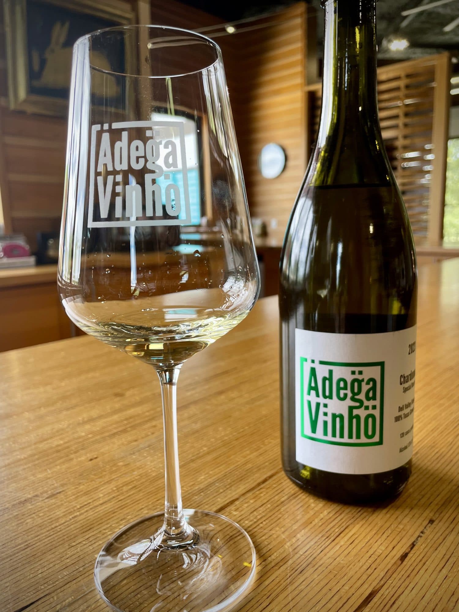 A glass of Adega Vinho estate Chardonnay beside its bottle on the wood tasting bar in Stonewall.