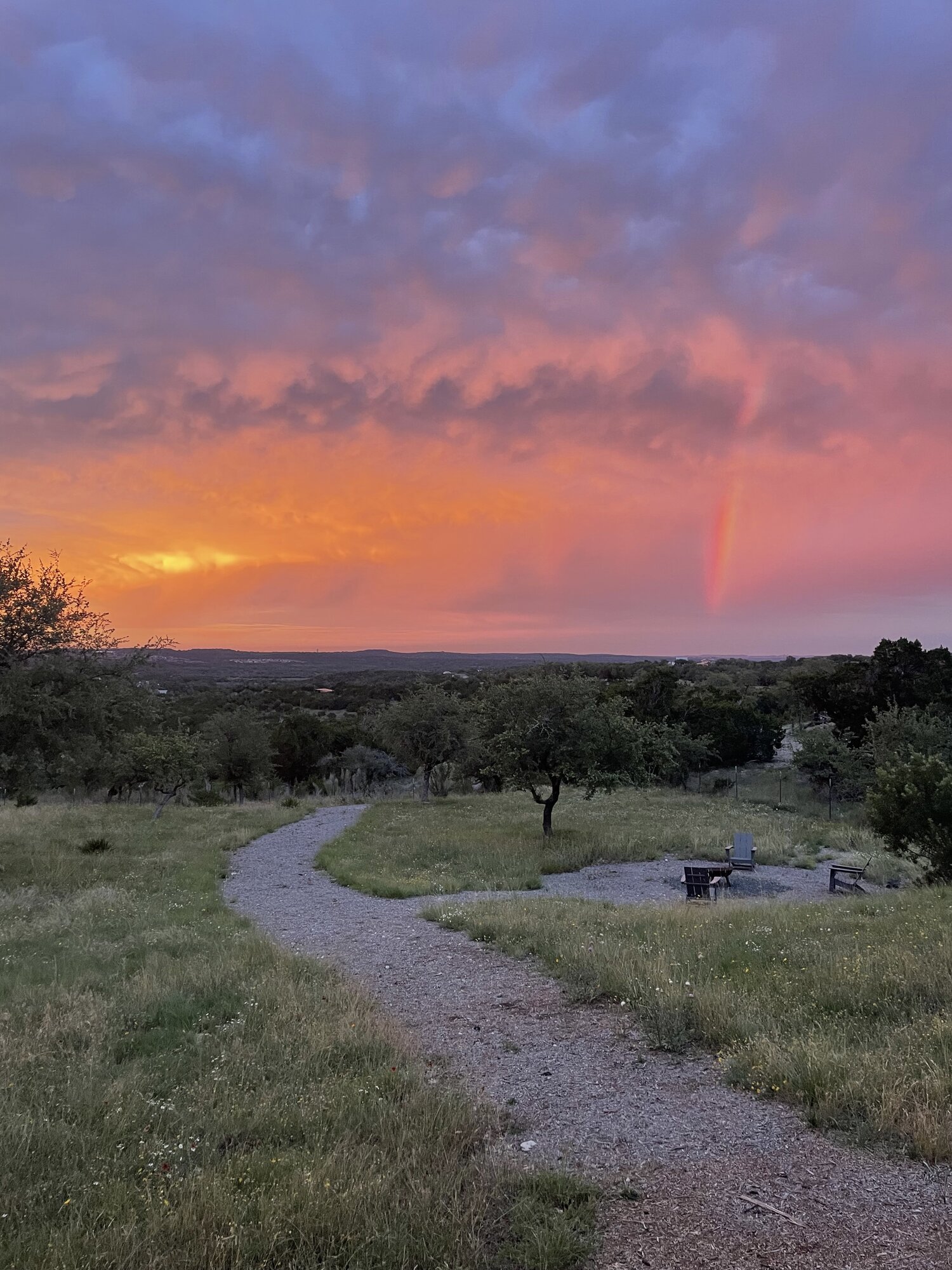 A Hill Country sunset and rainbow over oak trees, a gravel path, and chairs around a fire pit
